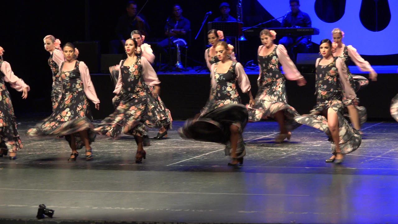 The dancers hold a wide formation on the Cabo San Lucas stage, with several faces visible and skirts swinging near the floor.