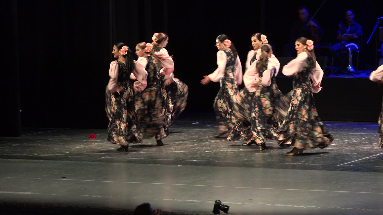 The dancers continue crossing the theater stage in Cabo San Lucas, Mexico, with long skirts lifted by movement and warm light separating them from the dark background.