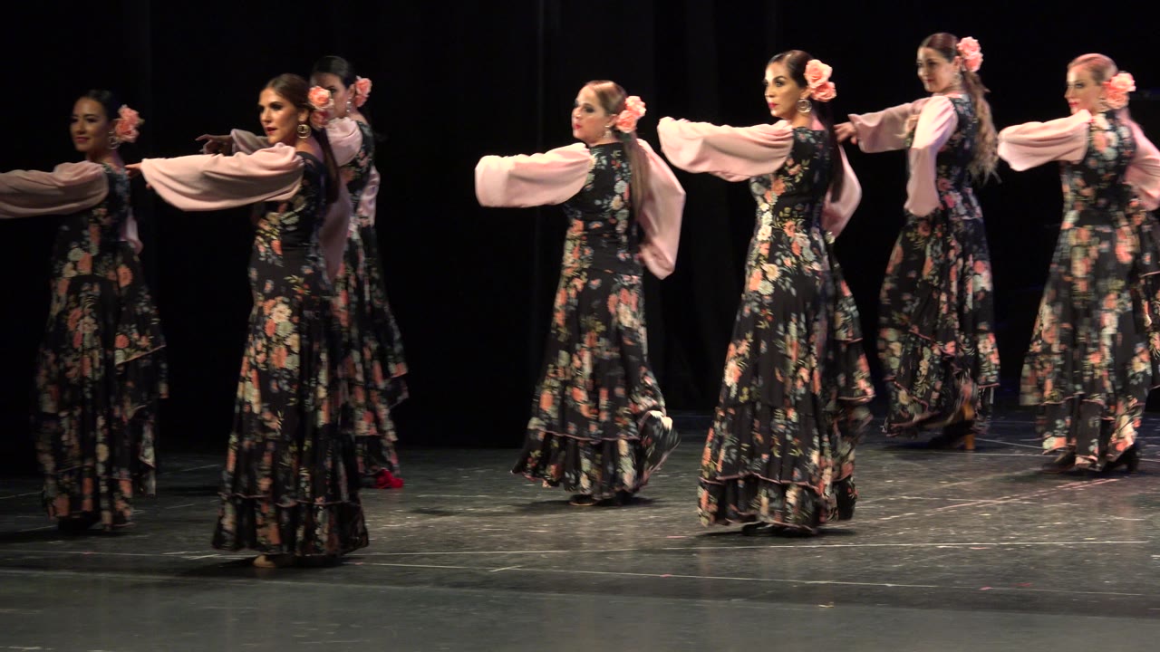 Women in floral dresses dance in a line on a dark stage.
