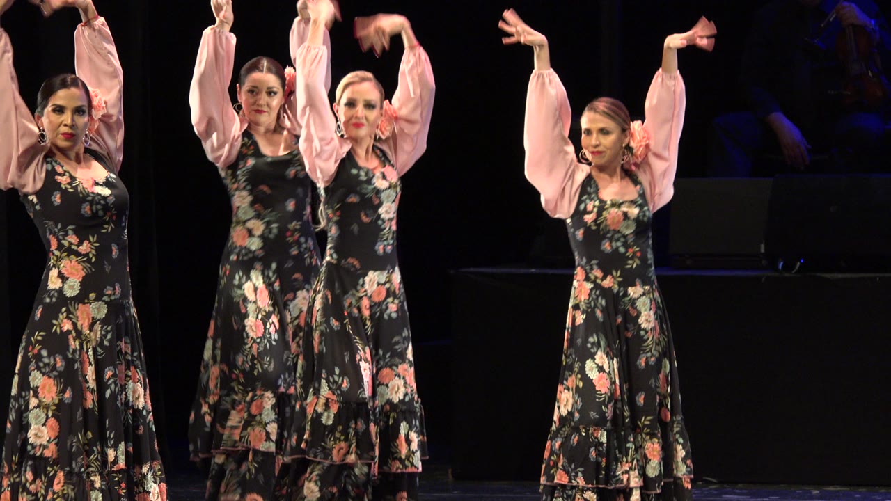 At 0:00, three adult women in floral dresses with pale pink sleeves raise their arms during a staged dance performance in Cabo San Lucas, Mexico.