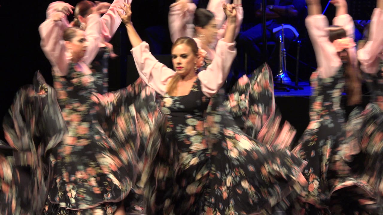 Women in matching floral performance dresses move across the stage in Cabo San Lucas, Mexico, with a front dancer facing the camera and other dancers blurred around her.
