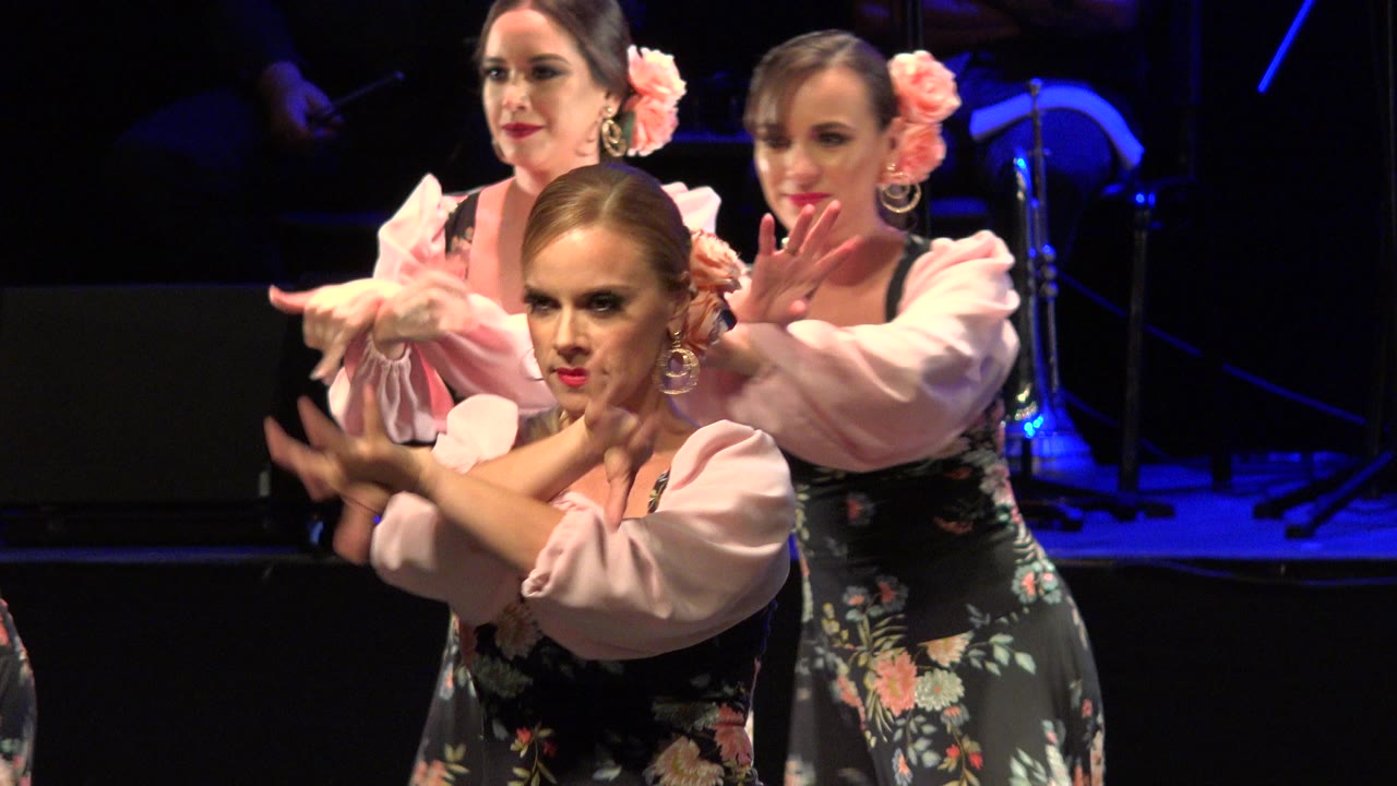 Three women in floral stage costumes perform hand gestures under blue stage light.