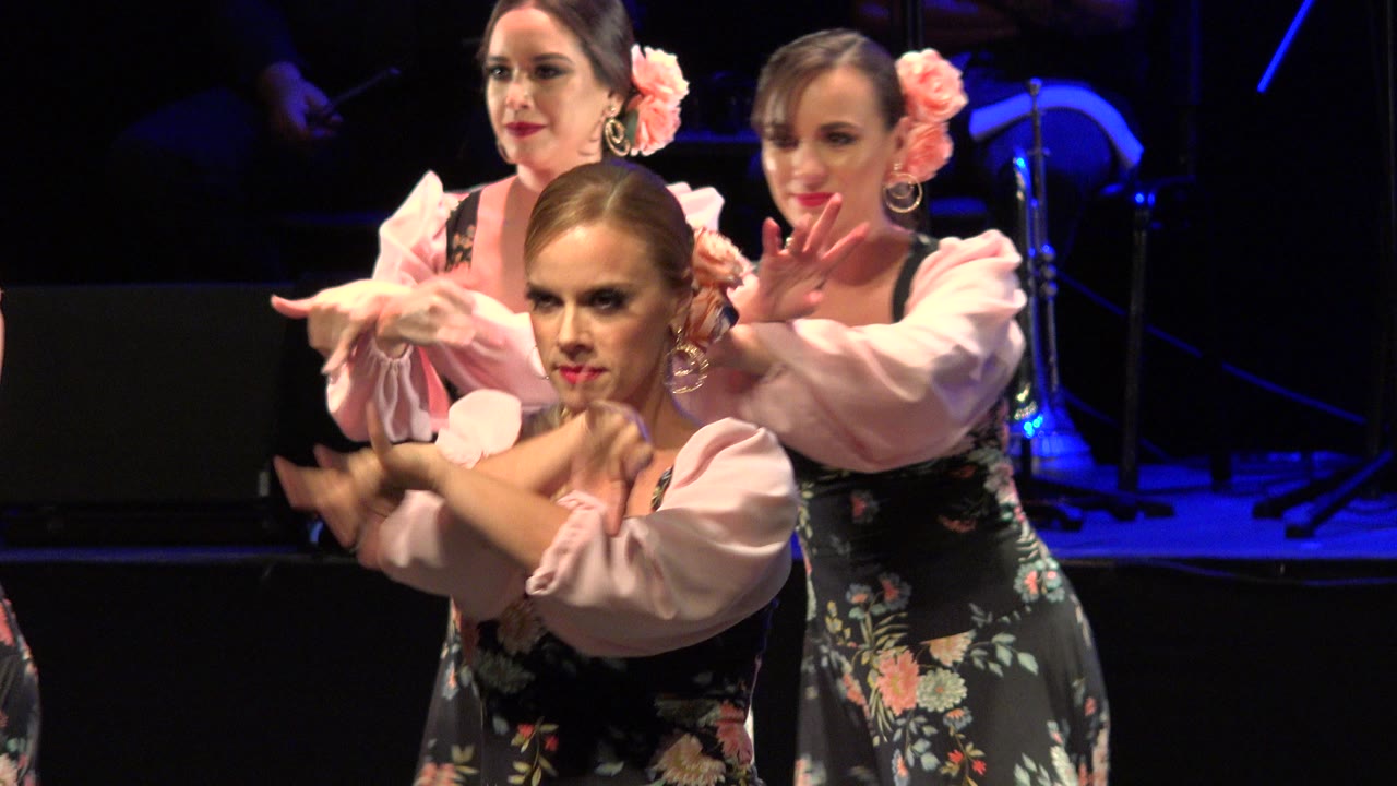Women in matching floral dresses continue the choreographed hand sequence, with faces visible and blue light behind them in Cabo San Lucas, Mexico.