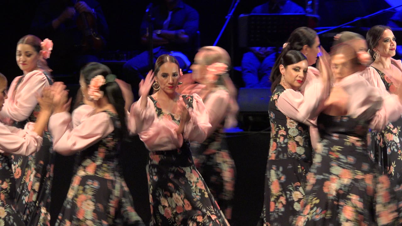 Women in floral dresses dancing close together on a blue-lit stage.