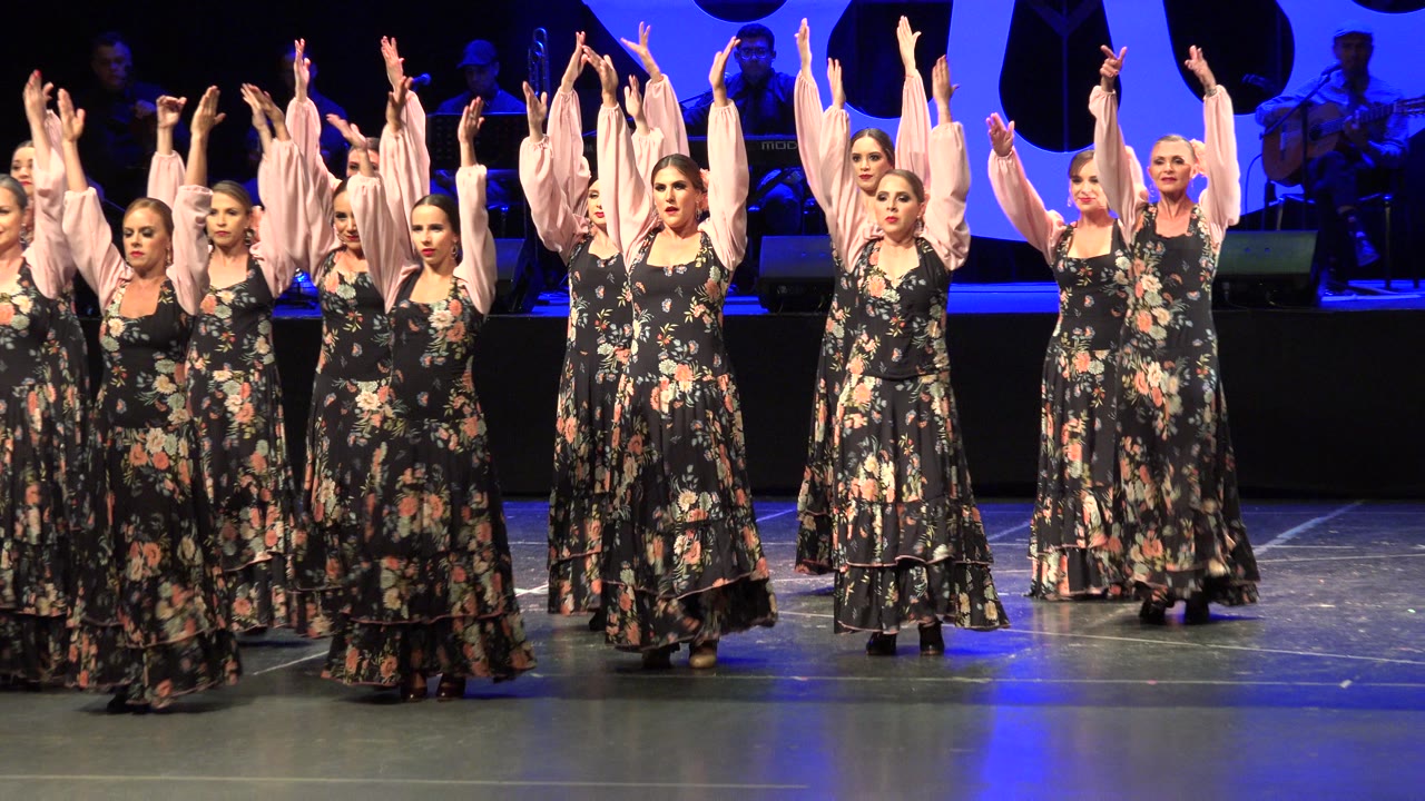 Adult women in black floral dresses with pale pink sleeves raise both arms during a stage dance performance in Cabo San Lucas, Mexico.