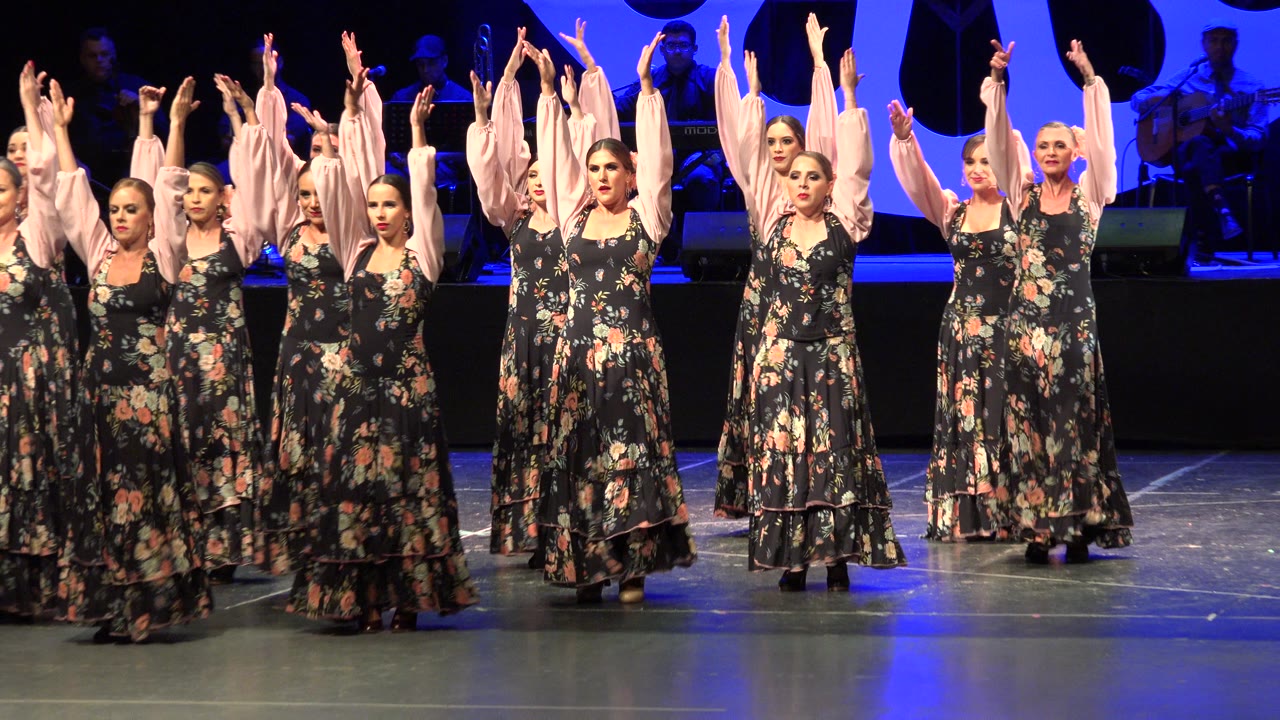 The dancers remain in formation with arms lifted, black floral skirts and pink sleeves catching warm front light against blue stage lighting in Cabo San Lucas, Mexico.