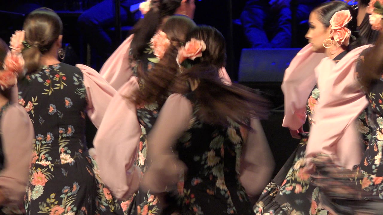 Women in black floral and pink dance dresses turn on a stage.