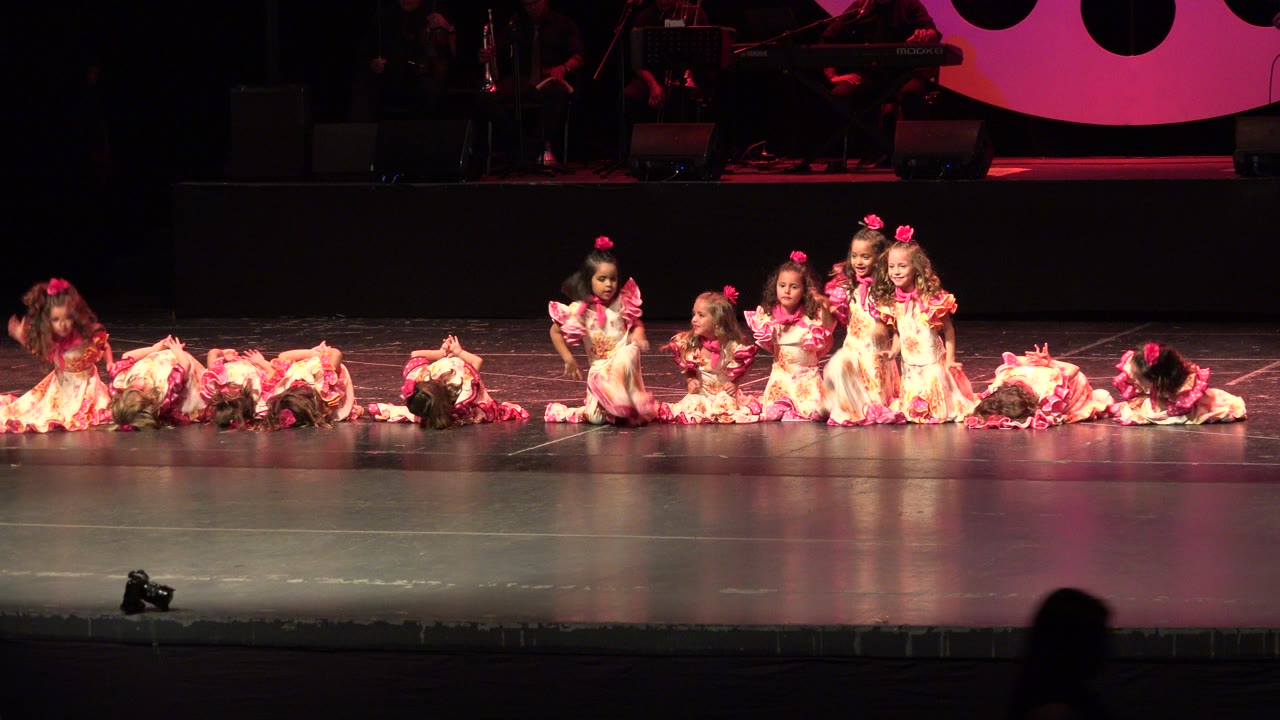 Young girls in ruffled dresses seated and kneeling on a stage.
