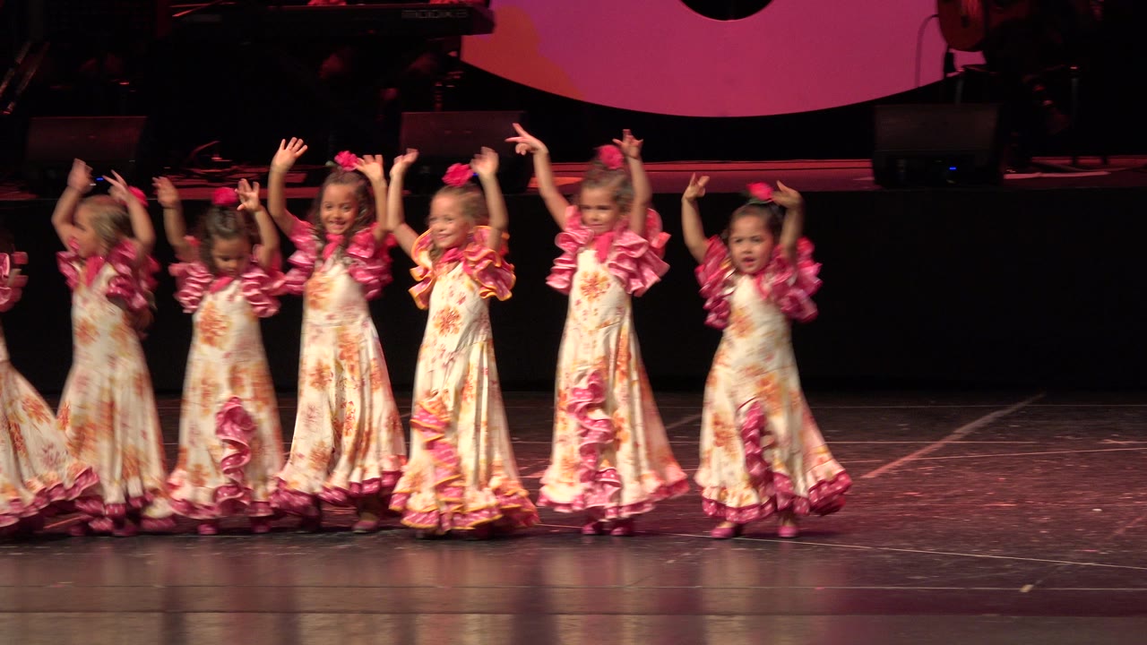 Young girls in floral dresses dancing on a stage with raised arms.