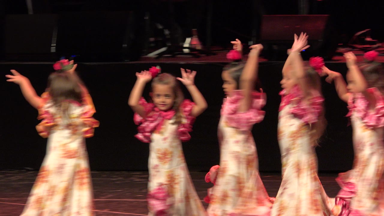 In Cabo San Lucas, Mexico, young girls in cream floral dresses with pink ruffled sleeves lift their arms during a stage dance routine.