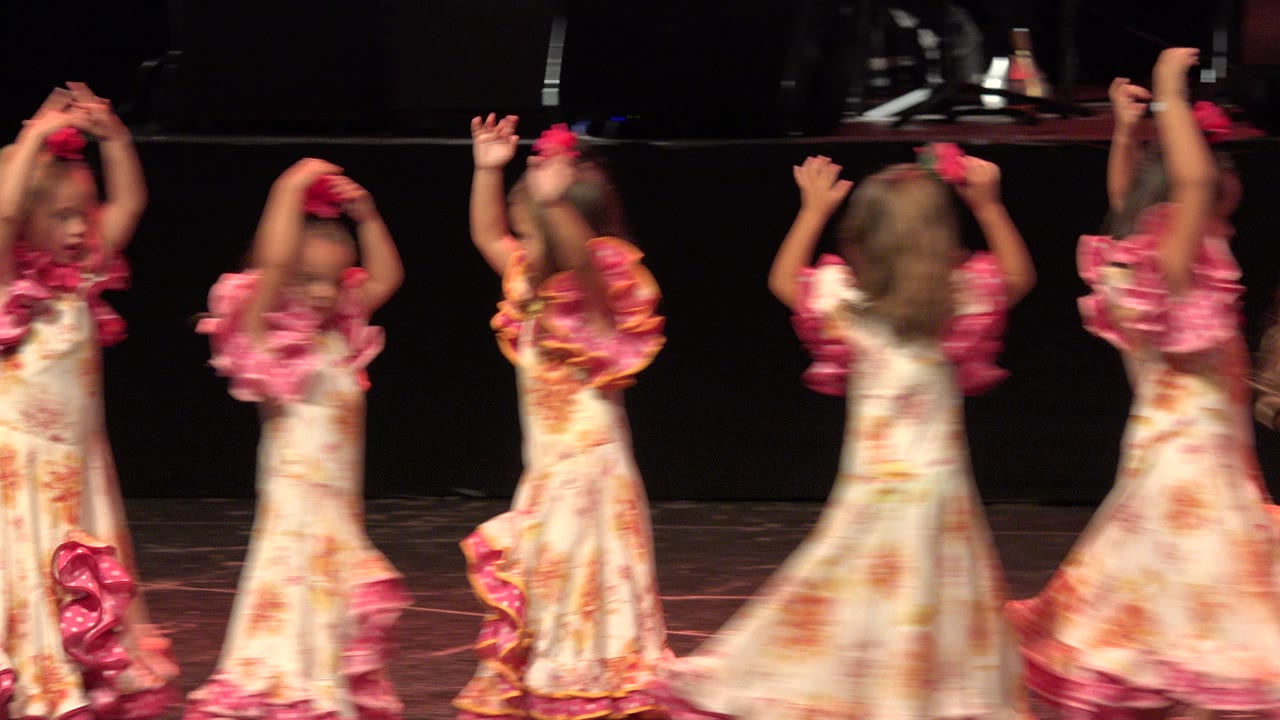 A cluster of young girls in floral dresses and bright pink ruffles perform arm movements on a dark theater stage in Cabo San Lucas, Mexico.