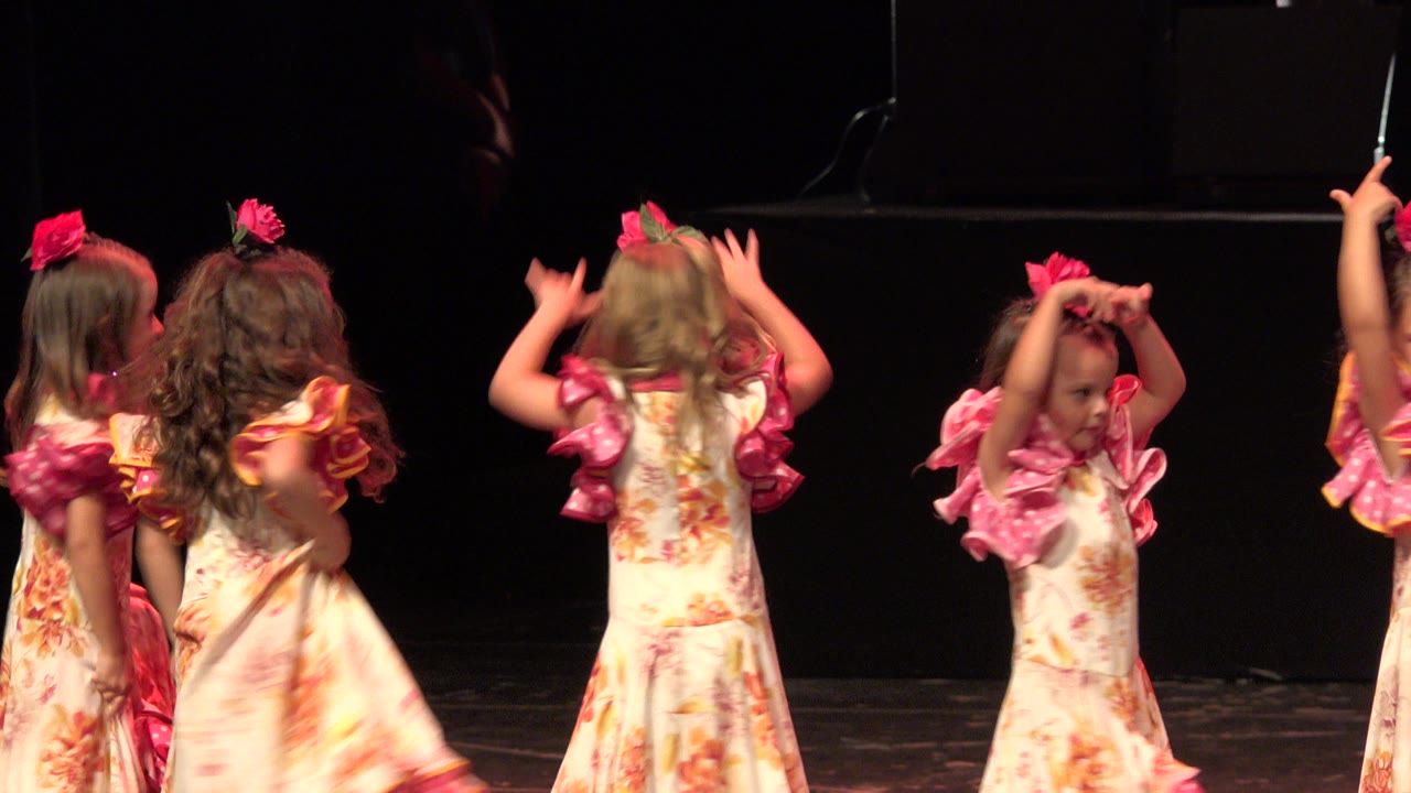 Several young girls in Cabo San Lucas wearing cream floral dresses with pink ruffled sleeves cluster on a dark stage as one child raises both hands near her head.