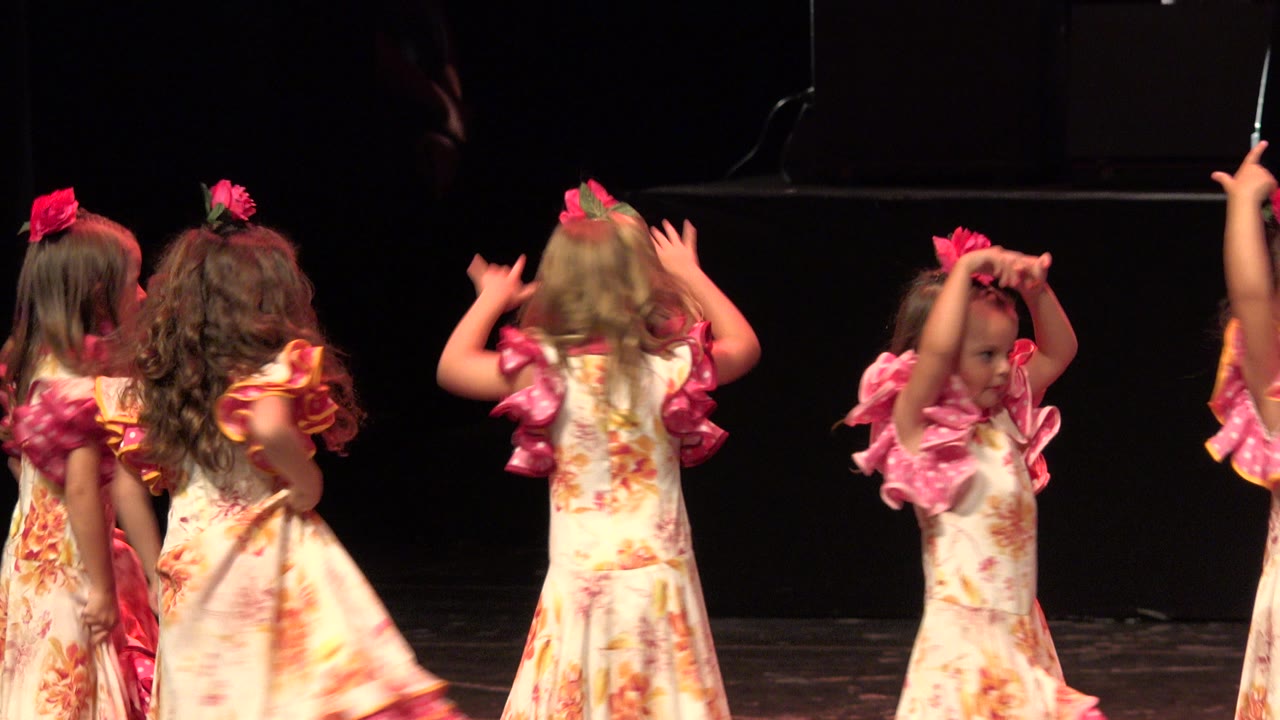 Young girls in Cabo San Lucas stand close together on a black stage, with one child turned away and another partially facing the camera in a pink bow and ruffled sleeves.