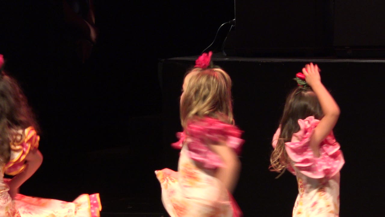 Two young dancers in pink ruffled costumes face away and lift their arms on the dark stage during the Cabo San Lucas recital.