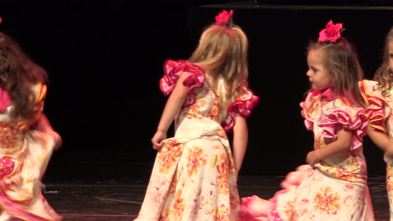 Young girls in floral pink ruffled costumes move across a dark indoor stage during a 2019 Cabo San Lucas dance performance.