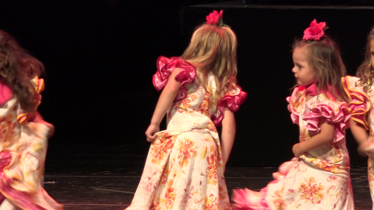 Three young girls in cream floral dresses with pink accents stand and turn on the stage during the Cabo San Lucas dance clip.
