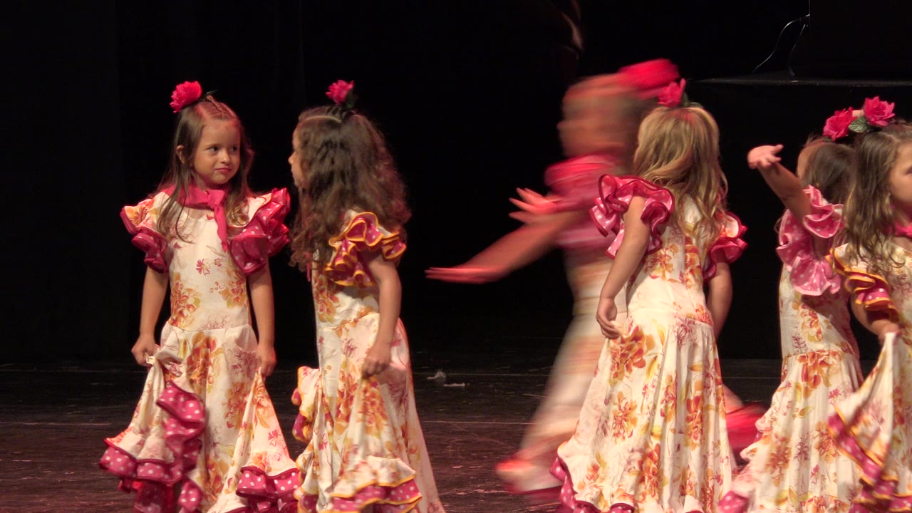 Young girls in floral dresses standing and dancing on a dark theater stage.