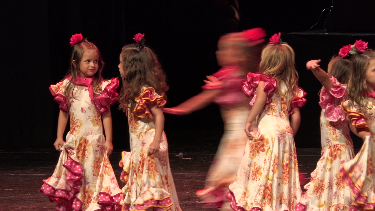 Girls in matching floral dresses dance across the Cabo San Lucas theater stage, with one child blurred as she crosses in front of the group.
