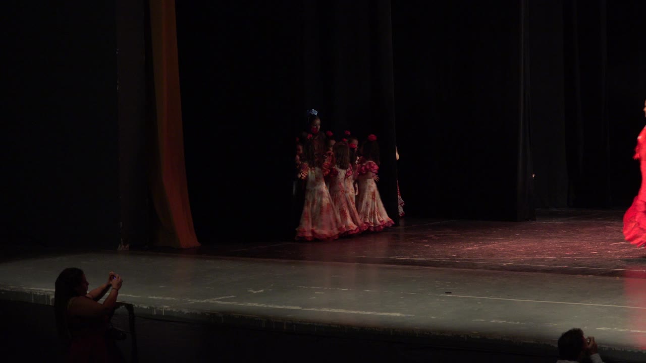 A wide audience view in Cabo San Lucas, Mexico shows a cluster of performers near the left curtain and a red dancer cropped at the far right of the stage.