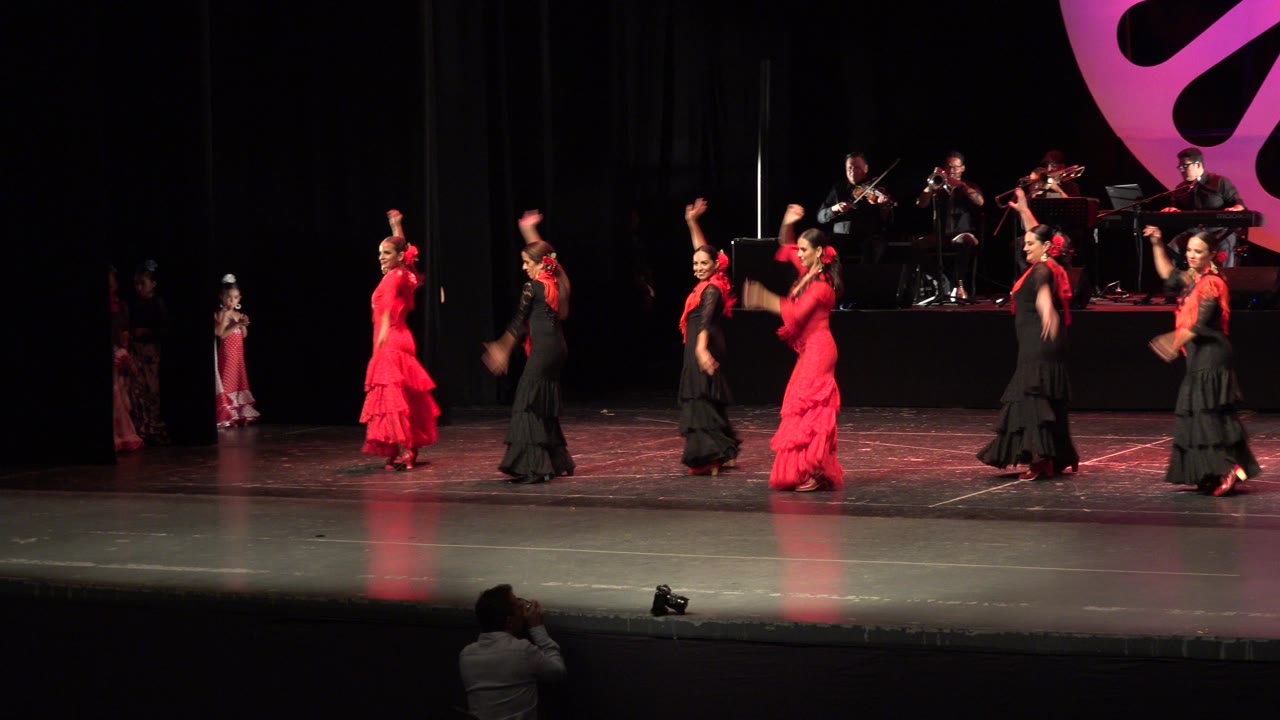 Dancers in red and black dresses perform on a dark theater stage with musicians behind them.