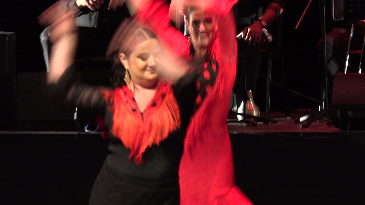In Cabo San Lucas, Mexico, the foreground dancer faces forward with blurred raised arms, red fringe across her black dress, and another performer and stage instruments visible behind her.
