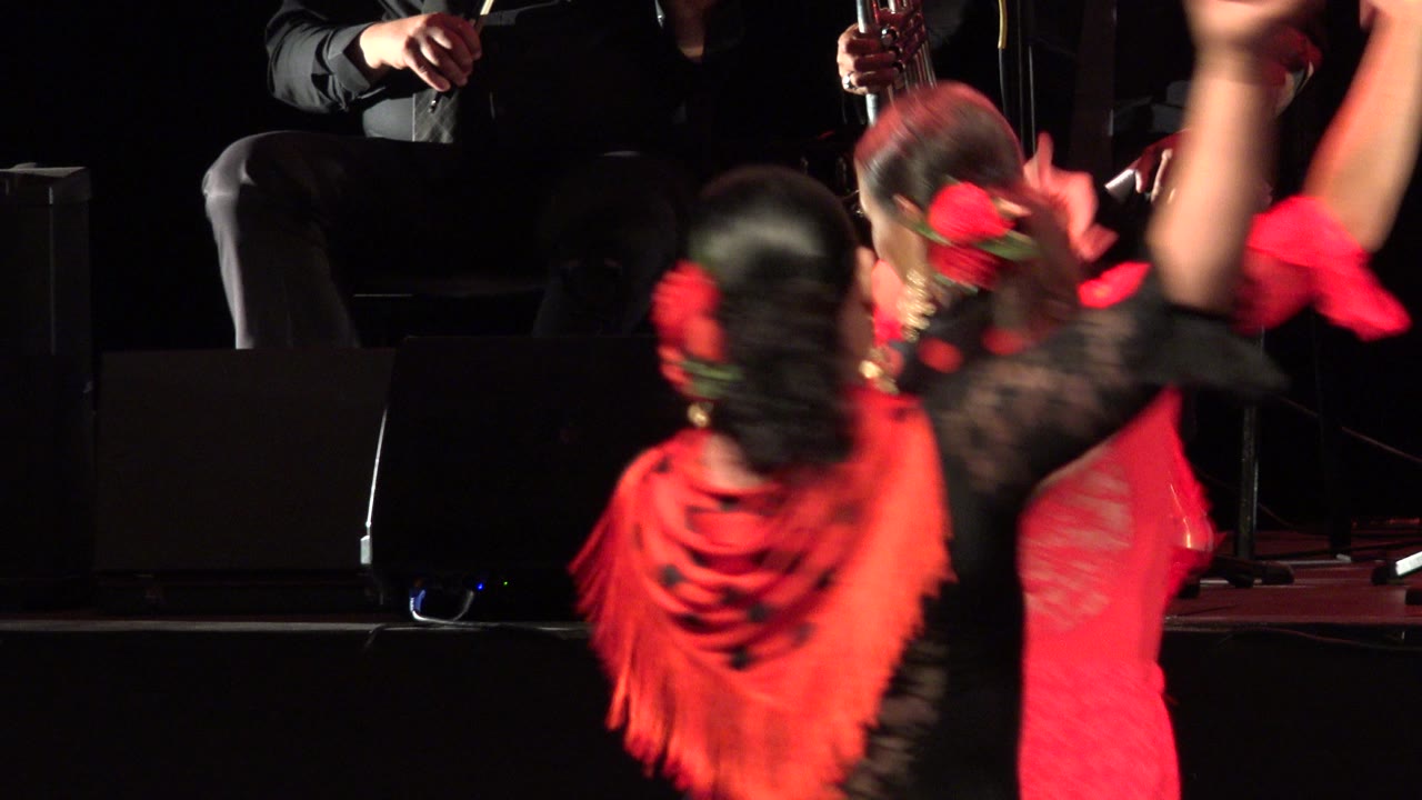 Two dancers in red and black costumes move in front of seated musicians on a dark stage.