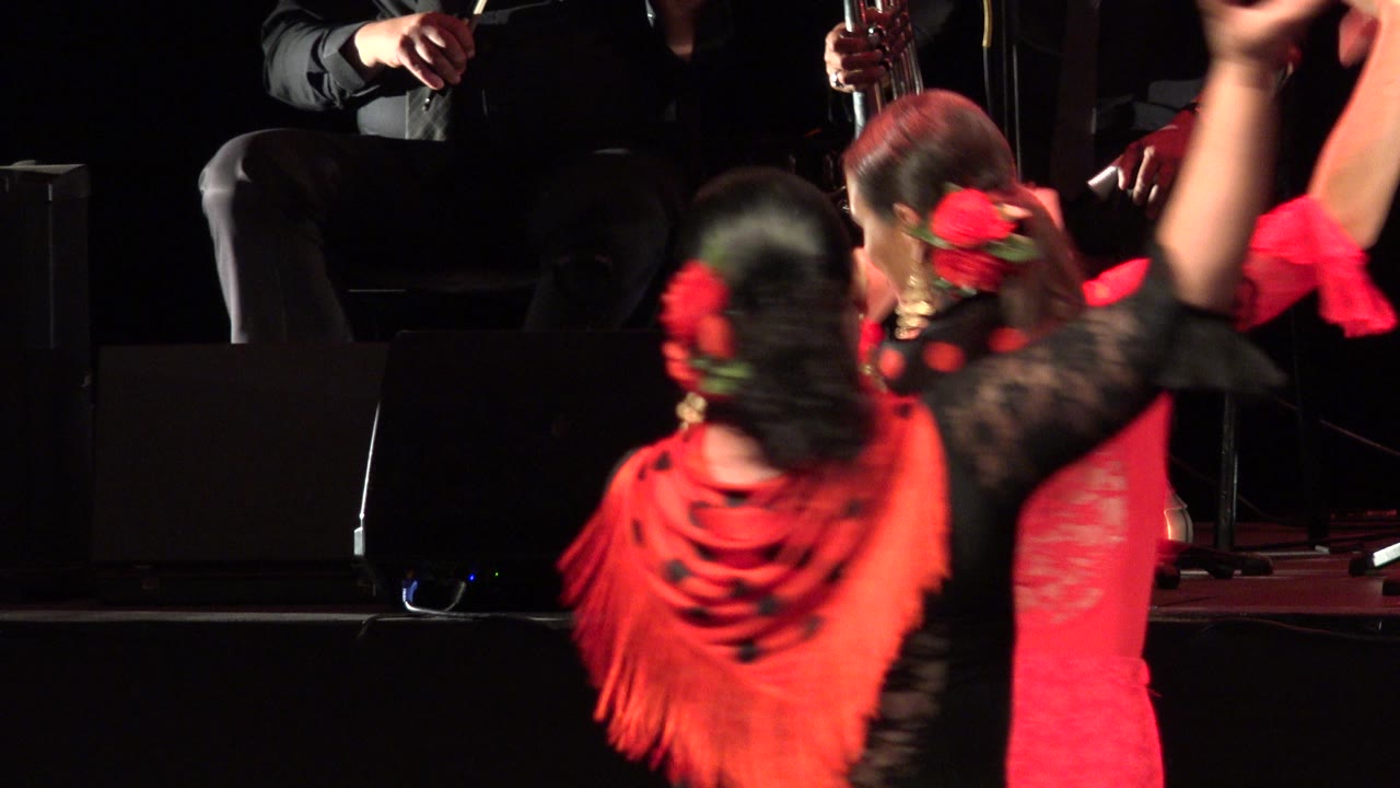 One second in, the Cabo San Lucas stage view shows dancers turned away and sideways, with red flowers, black lace sleeves, red fringe, and musicians seated in the rear.