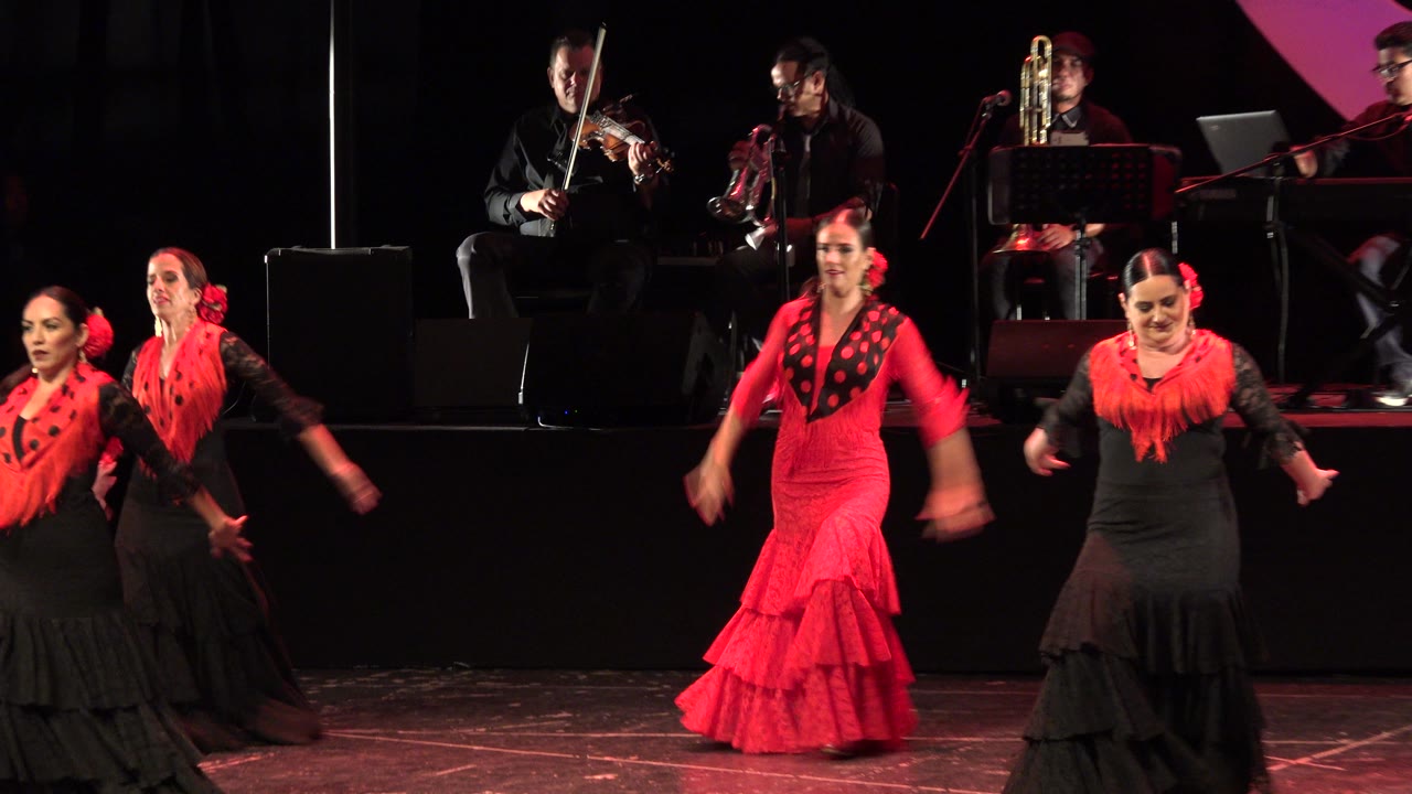 At 0:00 in Cabo San Lucas, women in red and black ruffled dresses perform on a dark stage while violin, horn, and keyboard players sit behind them.