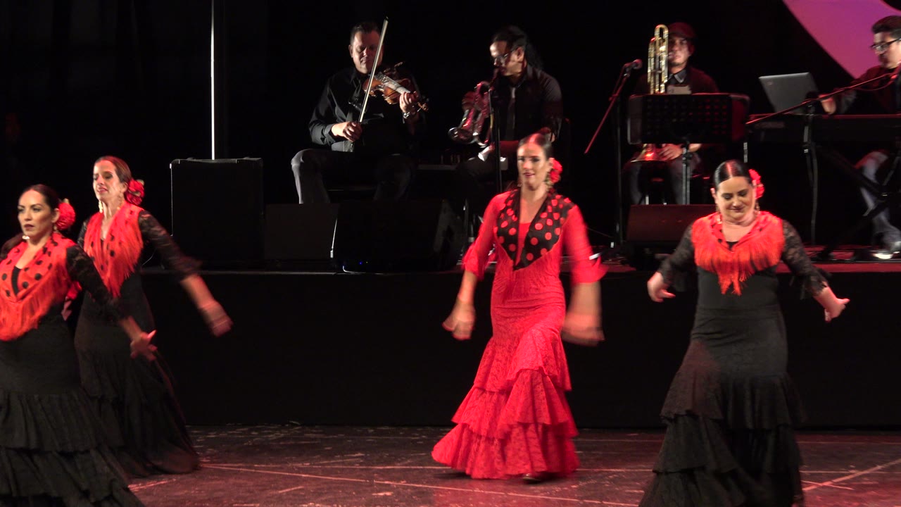 At 0:01 in Cabo San Lucas, the center dancer in a bright red ruffled dress steps forward while black-dressed dancers flank her and seated musicians remain in the background.