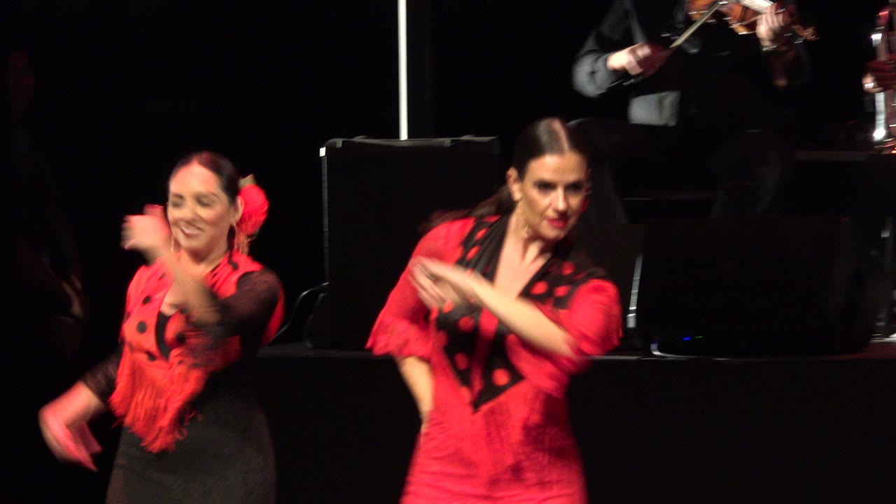 The two Cabo San Lucas performers continue synchronized arm gestures, with a large black stage monitor centered behind them and musicians cropped at the top edge.