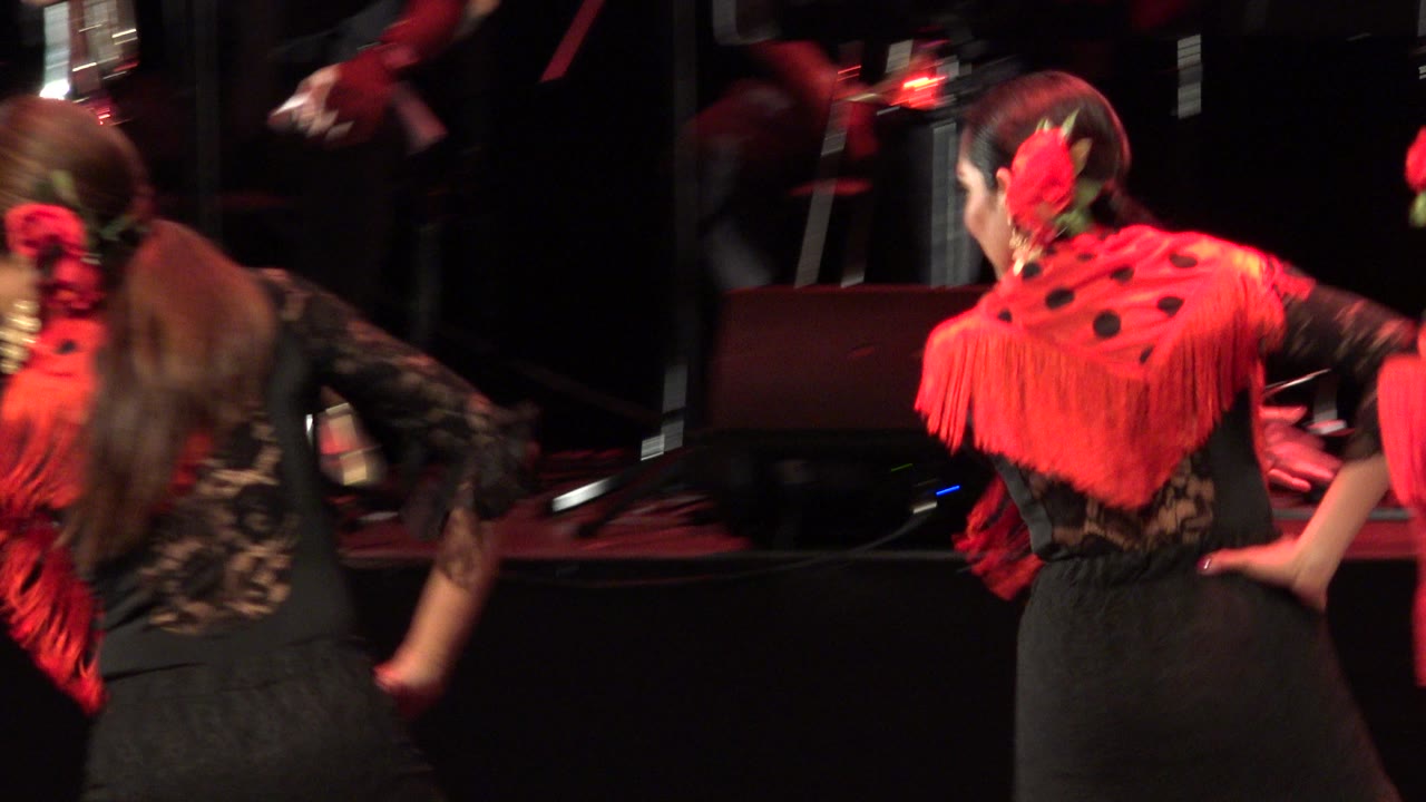 A red-shawled dancer turns away while another dancer crosses beside her during the Cabo San Lucas stage performance, with dark musician figures behind them.