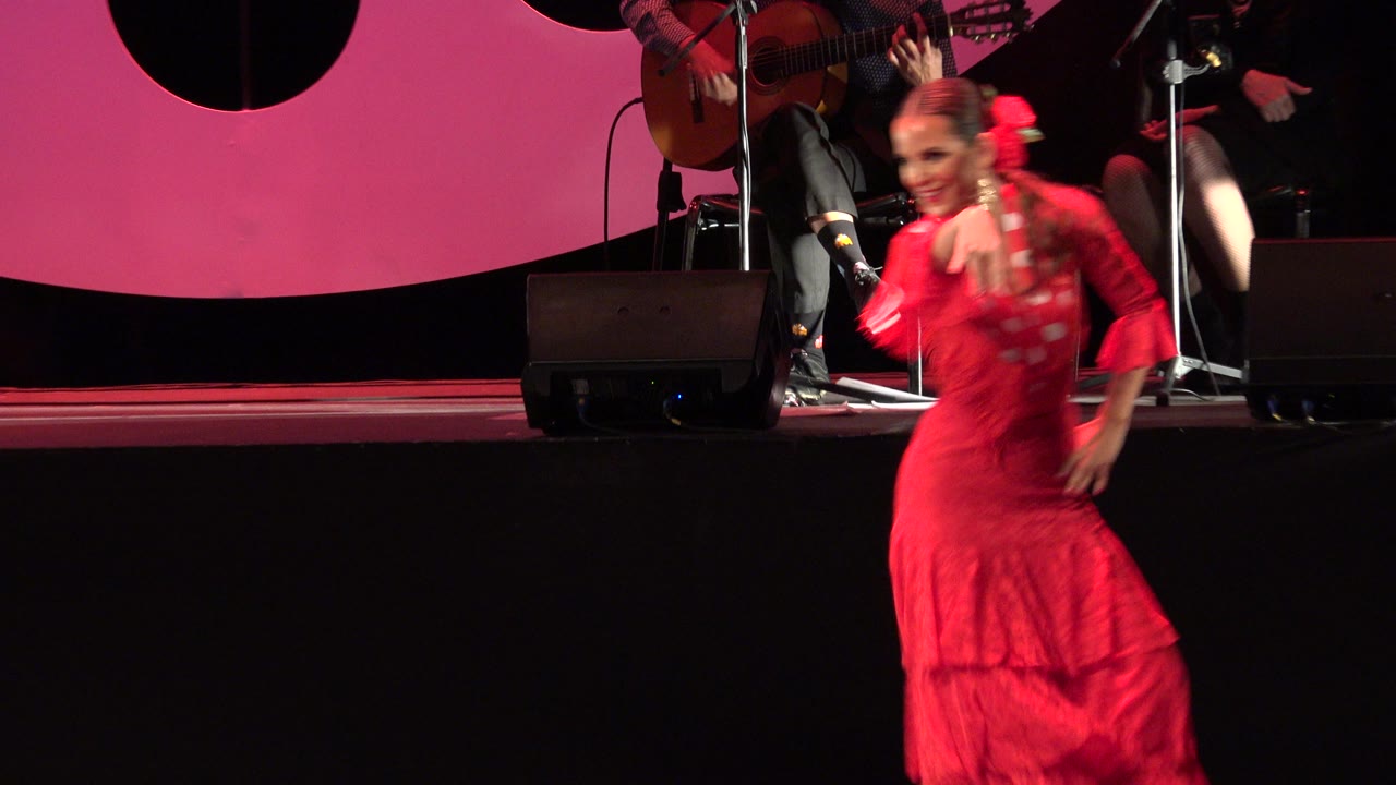 The red-dressed dancer remains close to the camera on the Cabo San Lucas stage while guitar players and equipment sit on the raised platform in the background.