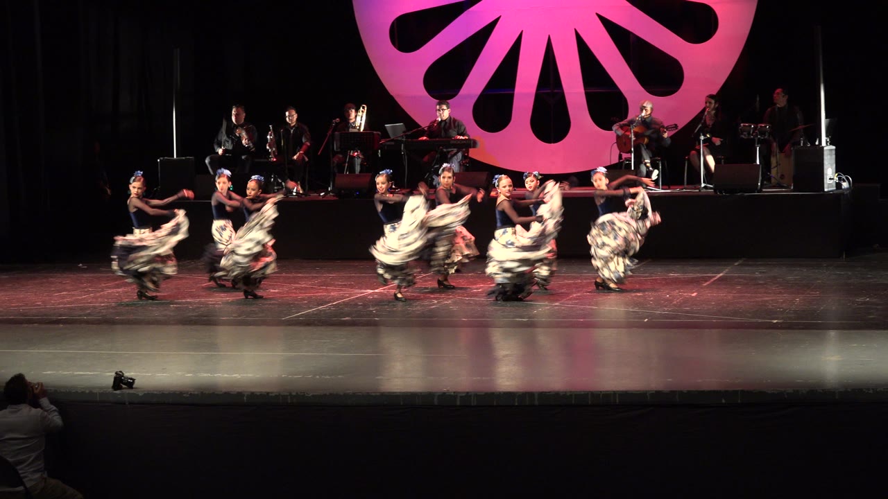 Dancers in patterned skirts perform on a theater stage with musicians behind them.