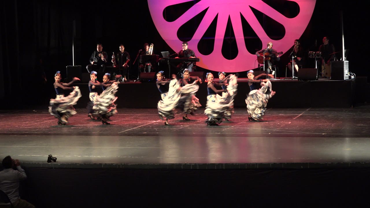 Dancers continue a coordinated skirt movement across the Cabo San Lucas stage, with the live band visible on the rear platform and warm pink lighting on the backdrop.