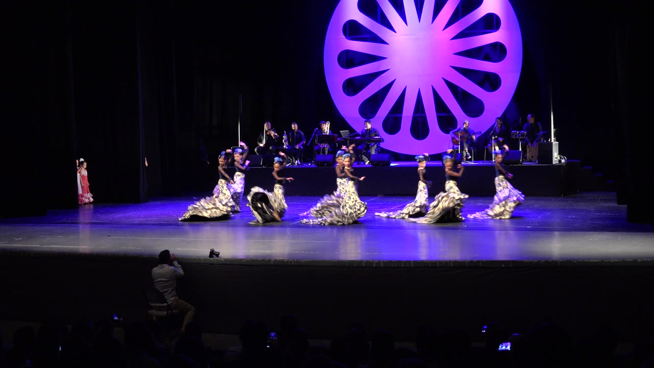 Cabo San Lucas stage performance with dancers clustered near center stage, a live band behind them, dark curtains, and a large purple backlit circular set piece.