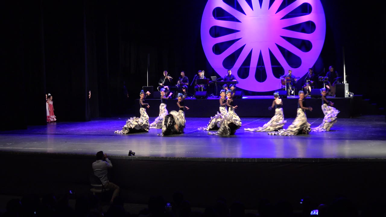 Wide Cabo San Lucas performance frame with dancers shifting into a broader formation, long patterned skirts catching blue-purple light, and musicians visible on the rear platform.