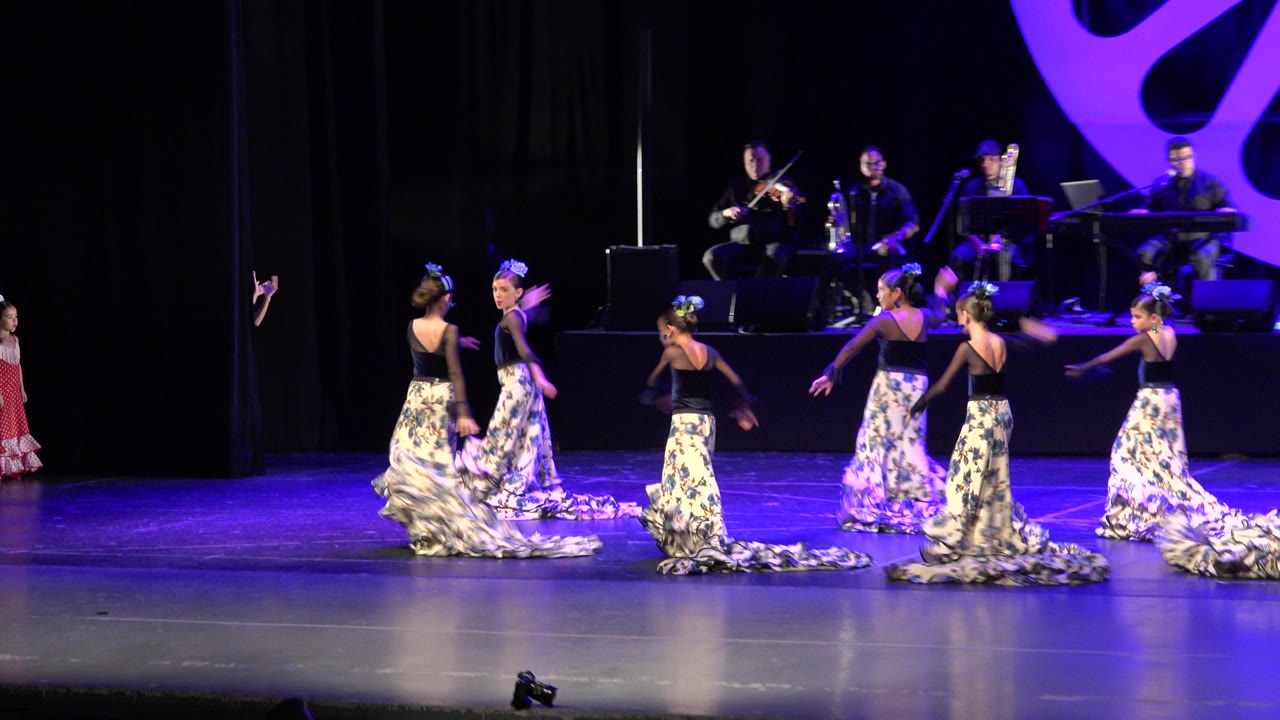 Young dancers perform on a theater stage with musicians behind them.