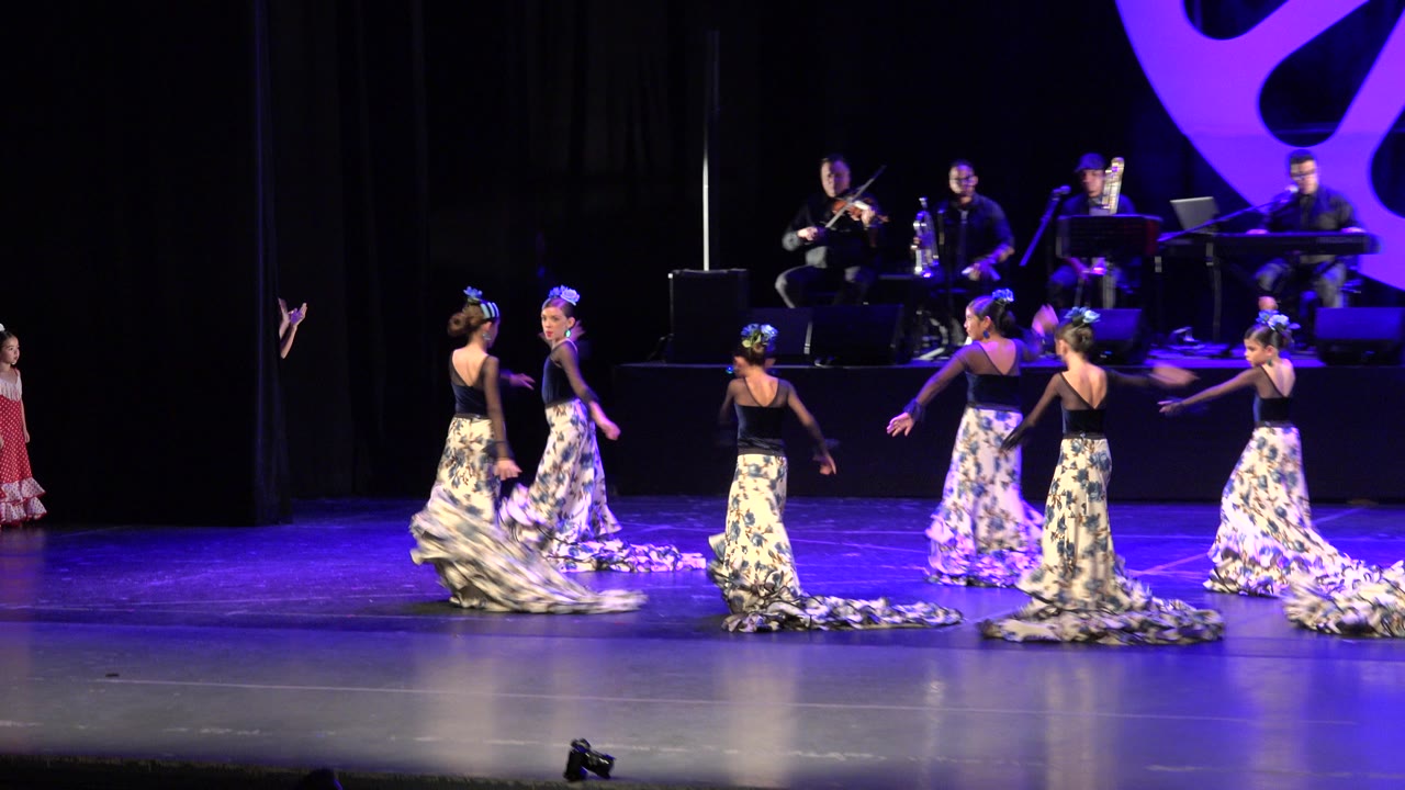 Several young dancers in black tops and long patterned skirts move across the Cabo San Lucas theater stage while musicians remain seated in the background.