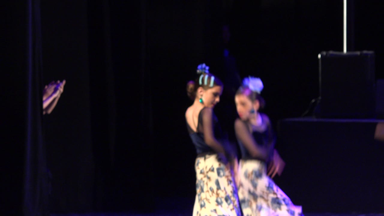 Two young girls in black tops and floral skirts dance on a dark stage in Cabo San Lucas, Mexico, with blue-purple light falling across their costumes.