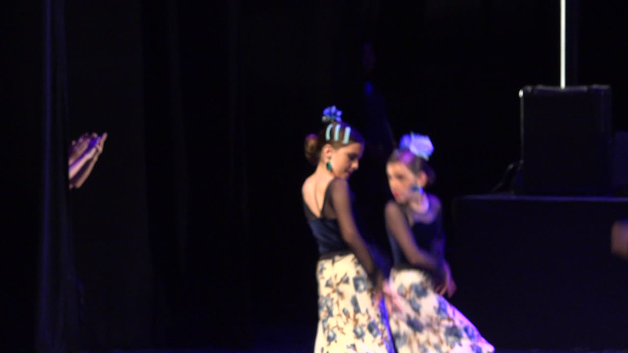 The two child dancers remain near center frame on the Cabo San Lucas stage, turning through the routine with a black equipment case visible behind them.