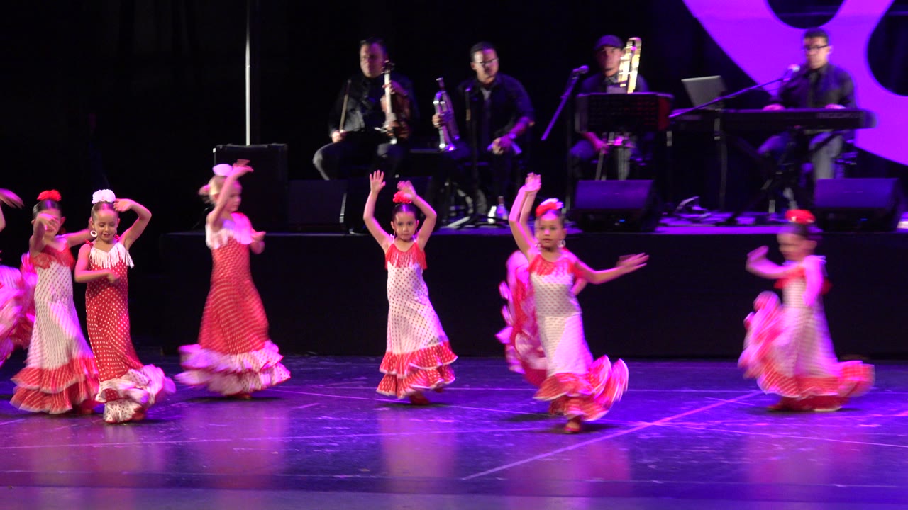 Young girls in red and white frilly gowns dance near the front of a stage in Cabo San Lucas, Mexico, with seated musicians and purple lighting behind them.