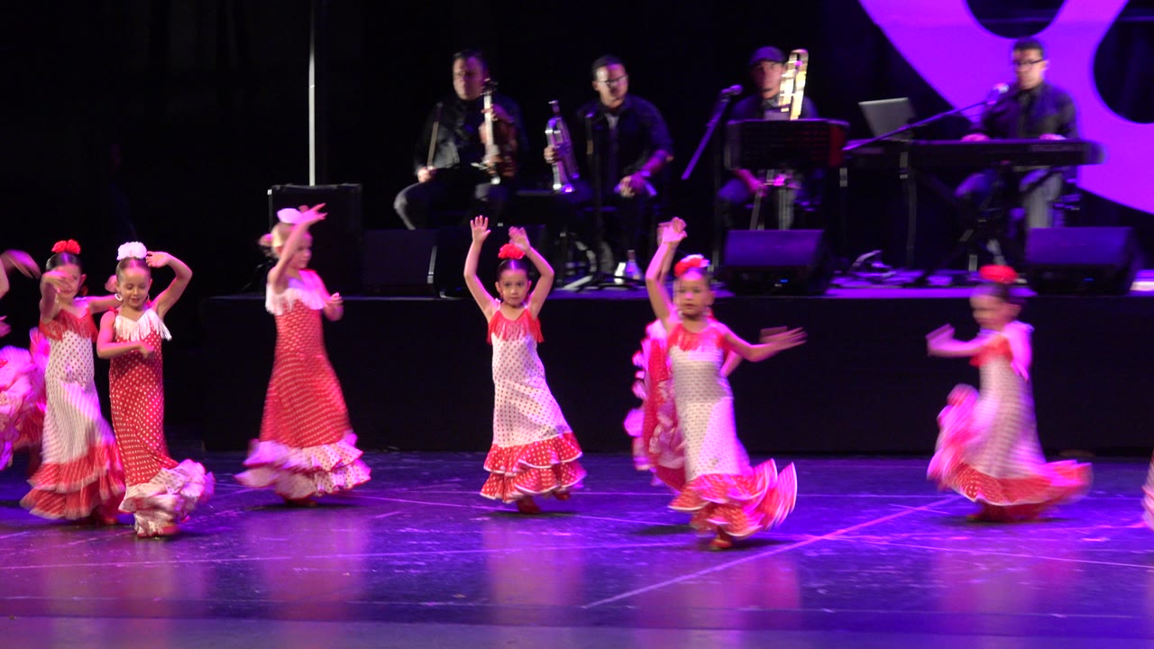 Children in coordinated red and white dresses move through a dance formation on a purple-lit stage in Cabo San Lucas, Mexico, while the band remains seated behind them.