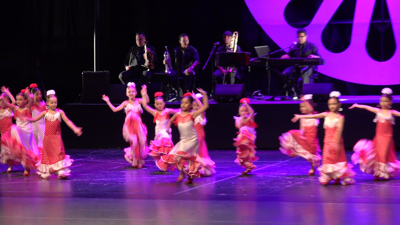 A group of girls in frilly dresses dance in front of a live band on a theater stage.