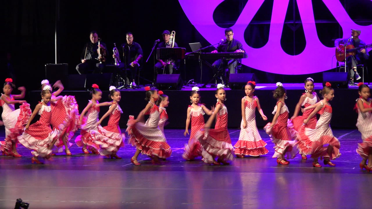 Girls in red and white frilled dresses dance on stage with musicians behind them.