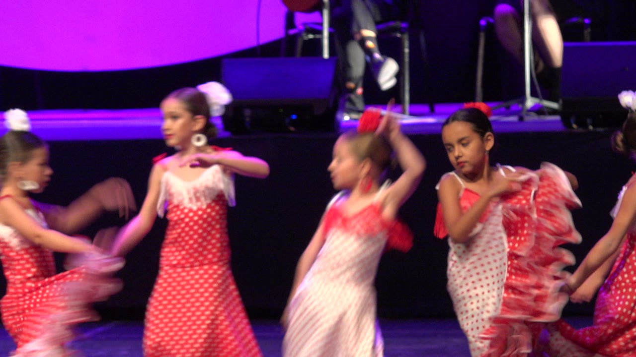 Several young dancers continue the routine in Cabo San Lucas, with one girl raising her arms while others move across the dark stage.