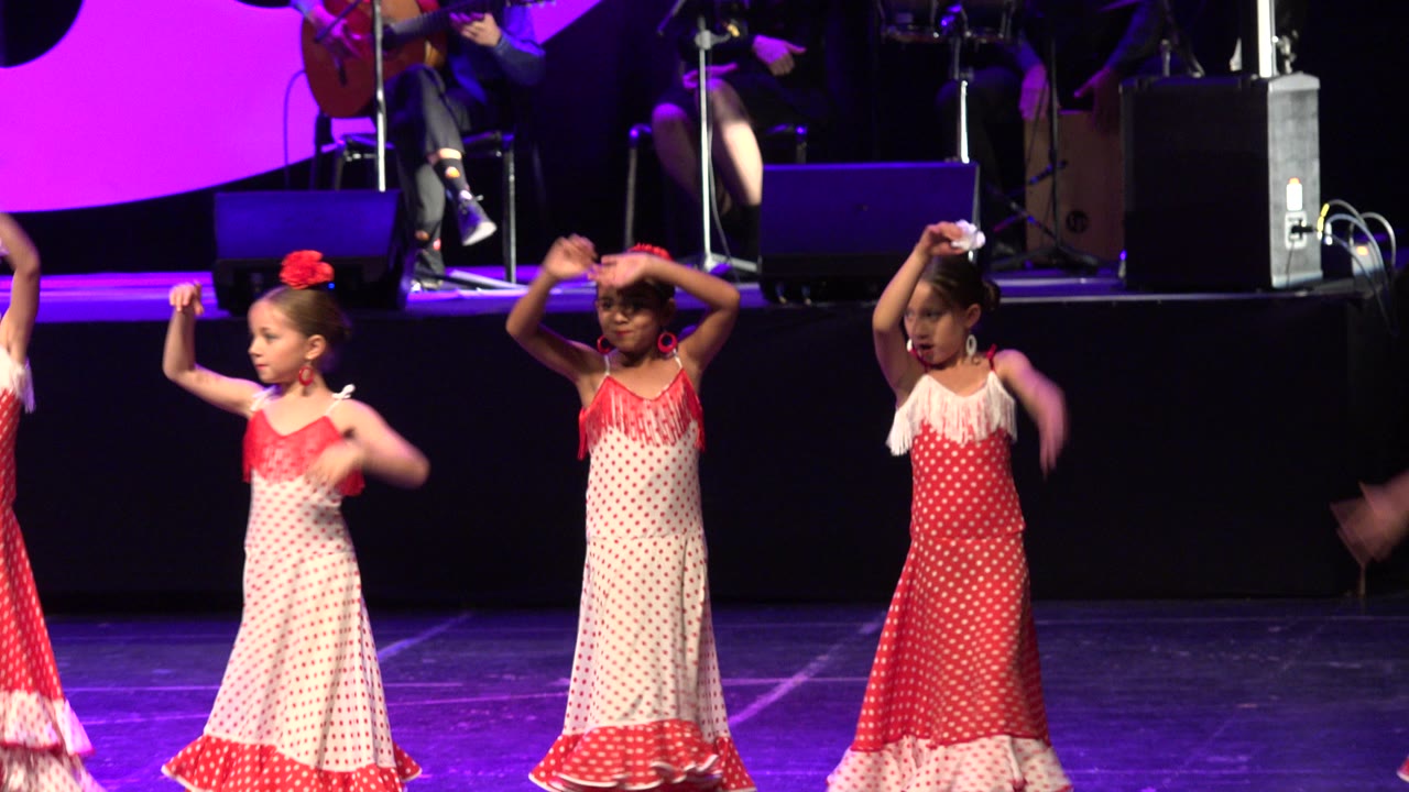 Three girls in alternating red and white dance dresses hold raised arm positions on the Cabo San Lucas stage while band equipment sits in the background.