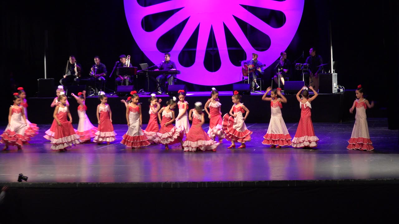 Children in pink dresses perform on a lit stage with musicians behind them.