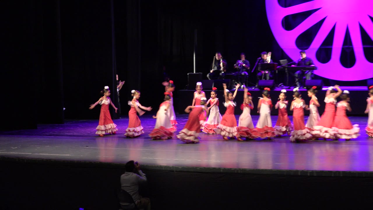 Children in ruffled red and white dresses continue across the Cabo San Lucas stage as the rear musicians remain seated beneath the magenta stage design.