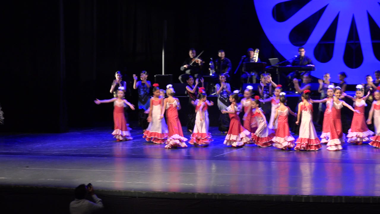 Young girls in white and pink dresses dance on a theater stage with musicians behind them.