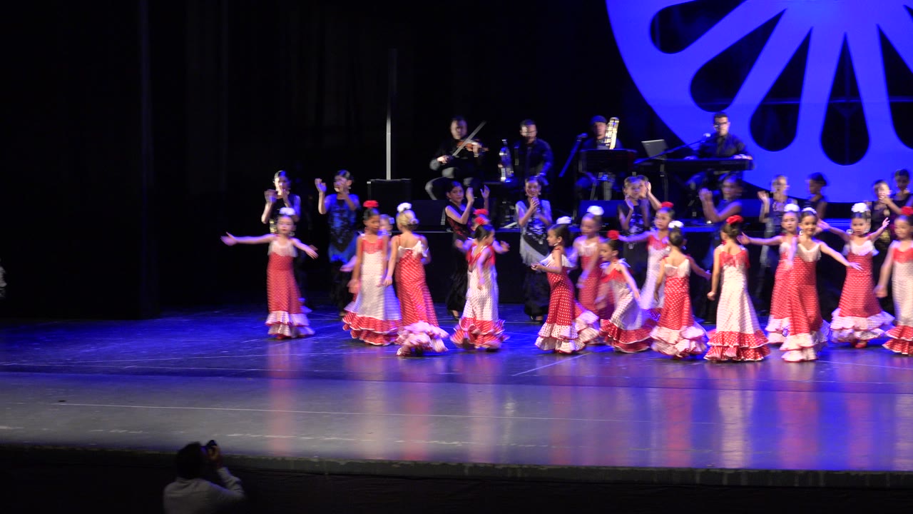 At 0:01 in Cabo San Lucas, the children remain grouped across the front of the stage, with red skirt trim catching the blue stage light and musicians visible behind them.